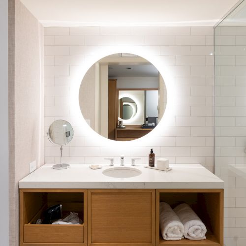 A bathroom vanity with a round backlit mirror, white countertop, wooden drawers, shelves, and neatly folded towels. 140 characters.