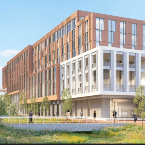 Modern multi-story building with brick and white-framed facade, glass windows, surrounding greenery, people walking along a landscaped lakeside path.