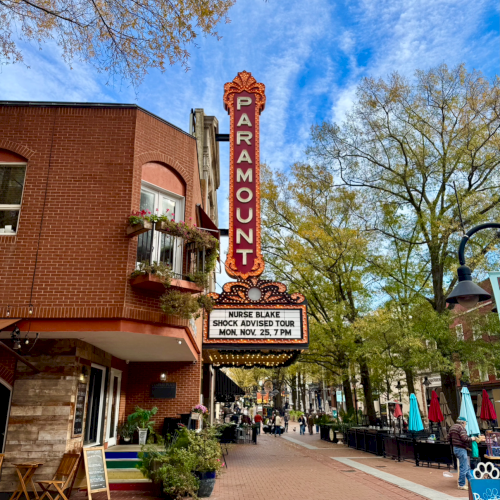 A sunny street scene with a brick building, cafe chairs outside, pedestrians, and a sign tower; trees line the sidewalk.