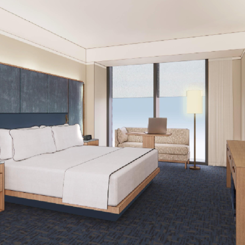 A modern hotel room with a king bed, blue upholstered headboard, nightstands, a seating area by the window, and a desk with a TV on the wall.