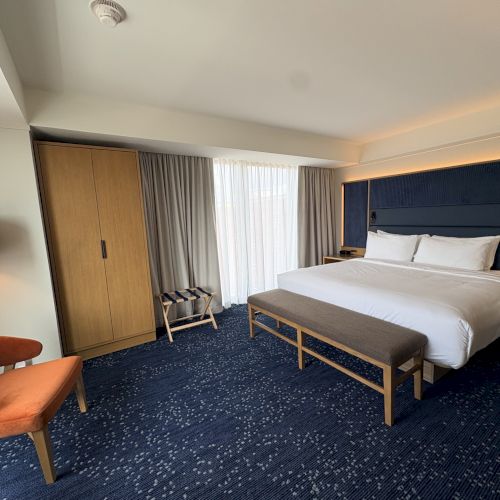A modern hotel room with a neatly made bed, curtains, wardrobe, lamps, and a view of the evening sky.