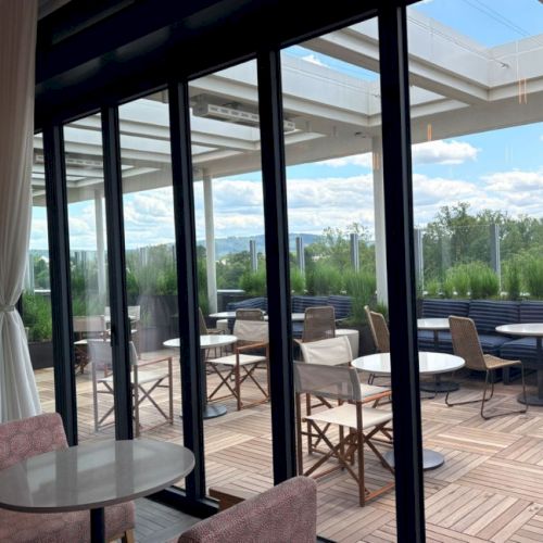 A modern patio dining area with round tables and chairs, viewed through large glass doors with trees and a skyline beyond.