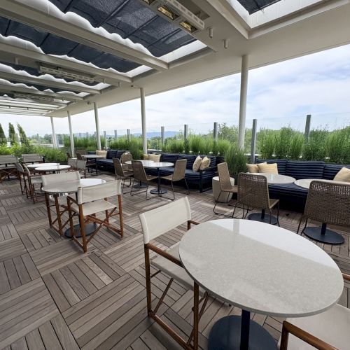 Covered outdoor patio with round tables, chairs, a blue sectional sofa, and greenery beyond the glass.