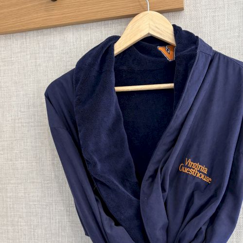A navy bathrobe with the &ldquo;Virginia Guesthouse&rdquo; logo hangs on a wooden hanger against a wall.