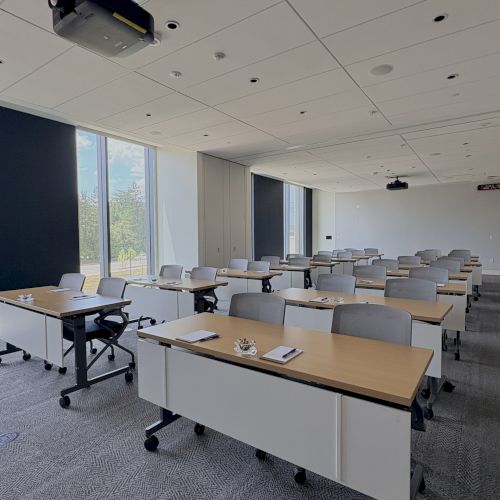 A modern classroom/conference room with rows of desks, gray chairs, and a ceiling projector near large windows.