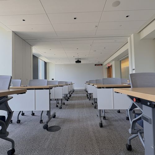 A bright conference room with rows of wheeled desks and chairs, a ceiling projector screen, and recessed lights in a clean office hallway.