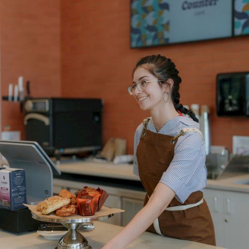A smiling barista in an apron serves at a caf&eacute; counter with pastries and a coffee pot while screens glow behind her.