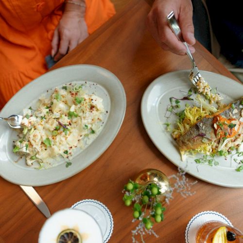 Two people serve gourmet dishes from a wooden table, plating creamy pasta and fish with herbs under warm light.