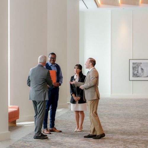 In a bright office lobby, four professionals in suits chat and greet while one holds a folder near framed photos.