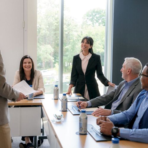 A business meeting in a conference room where a man hands papers to colleagues as a woman stands smiling beside the table.