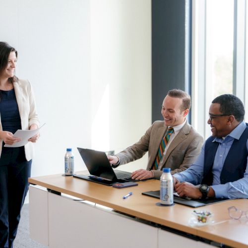 A business meeting: a woman presents papers while coworkers smile and work on laptops at a conference table.