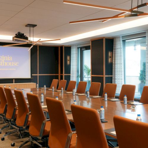 A modern meeting room with orange chairs, a long table, and a screen showing &ldquo;Virginia Guesthouse.&rdquo;