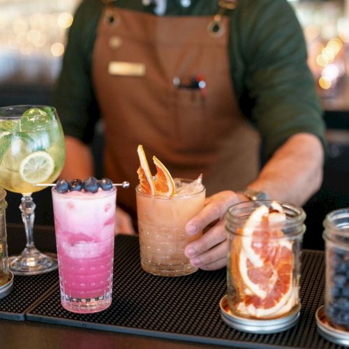 A bartender in an apron serves a citrus cocktail while other berry and lemon drinks sit on the bar in jars.