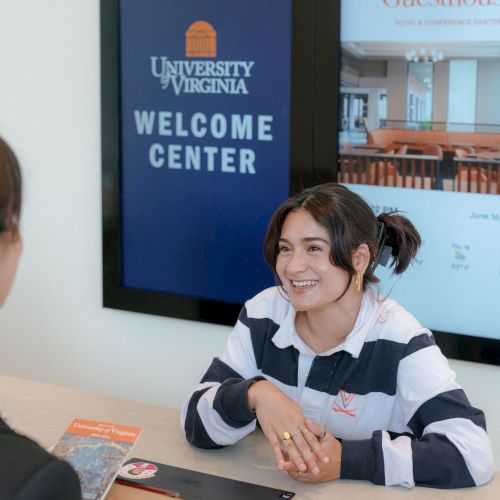 A smiling woman at a table during a University of Virginia Welcome Center visit, with screens showing UVA signage.