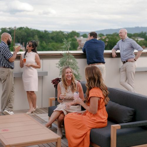 A rooftop gathering: people chatting and toasting with drinks on a sunny terrace, with city views behind them.