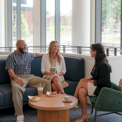 Three friends chat on a couch in a bright lounge, sipping drinks and eating snacks at a small round table.