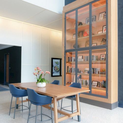 A modern office lounge with a wooden table, blue chairs, and a glass display case of books inside.