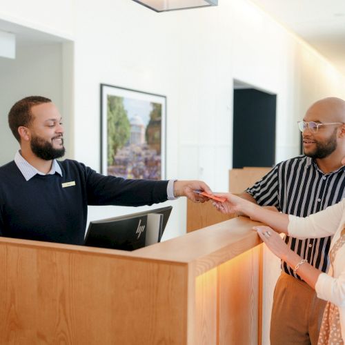 In a modern office, a staff member hands a visitor a card while coworkers chat at the reception desk.