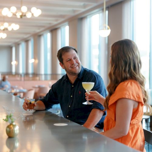 Two people chat at a bright bar, clinking glasses and holding drinks in a restaurant with warm lights.