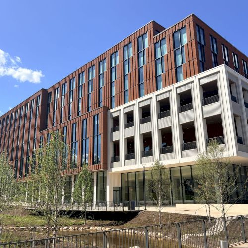 A modern multi-story building with red brick and white balconies, large windows, and trees in front under a clear blue sky.
