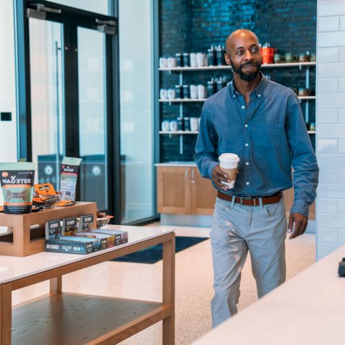 A man in an office lobby holds a coffee cup while standing by a table stocked with packaged snacks and drinks.