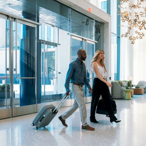 In a modern hotel lobby, a man and woman pull luggage carts while talking beneath hanging lights.
