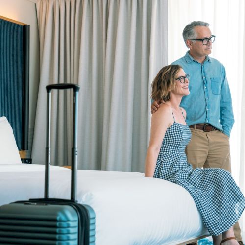 A smiling older couple stands by a hotel bed by curtains with a suitcase, the man gently holding the woman&rsquo;s shoulder.