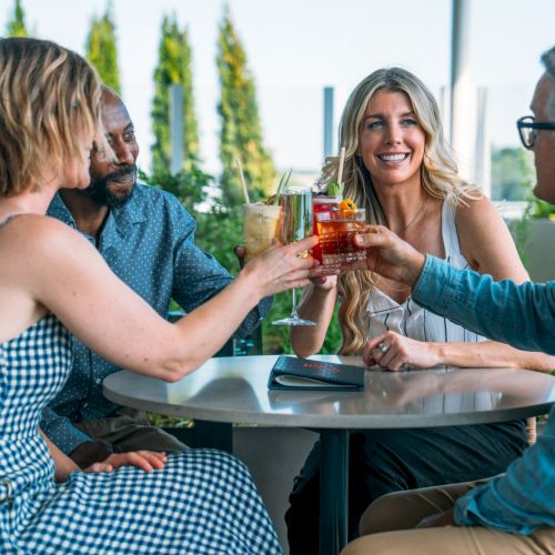 Friends clink glasses with colorful cocktails on a sunny outdoor patio, smiling and chatting together.