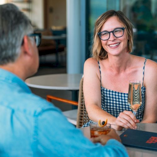 Outdoor caf&eacute;: a smiling woman with blue glasses chats with a man across the table, holding a drink.