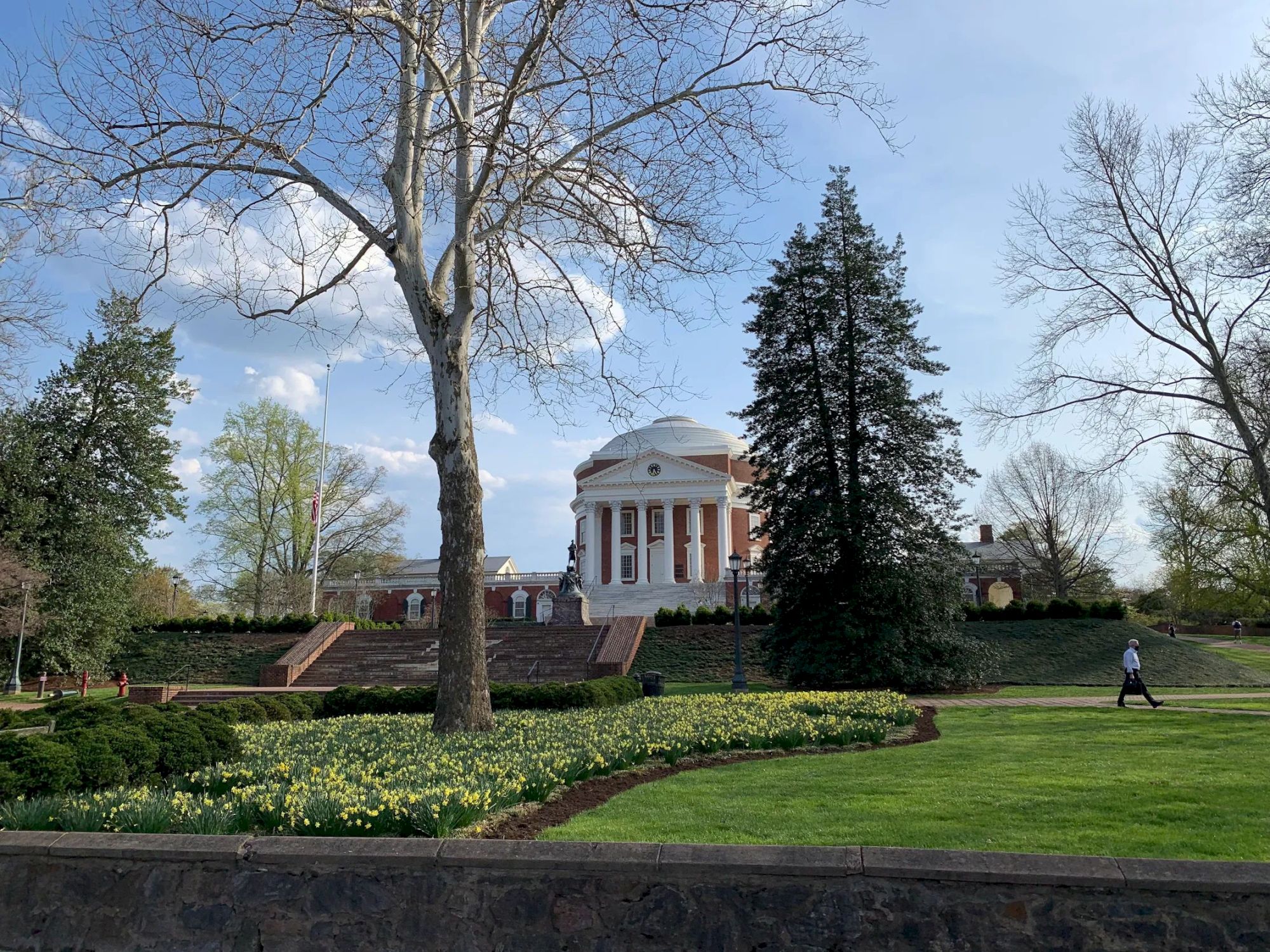 A classical domed white pavilion sits behind a trimmed lawn and flower beds, framed by tall trees on a sunny day.