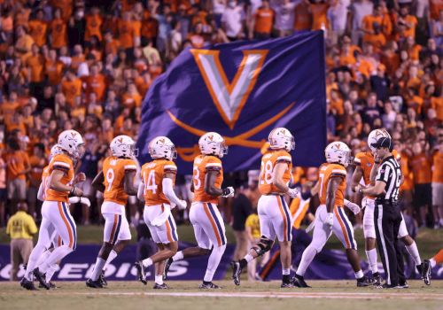 A college football team in orange and white uniforms lines up on the field as a large Virginia banner looms in the background, cheering fans abound.