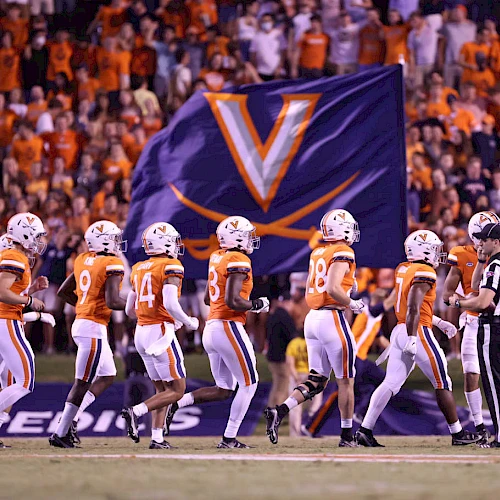 A college football team in orange and white uniforms lines up on the field as a large Virginia banner looms in the background, cheering fans abound.