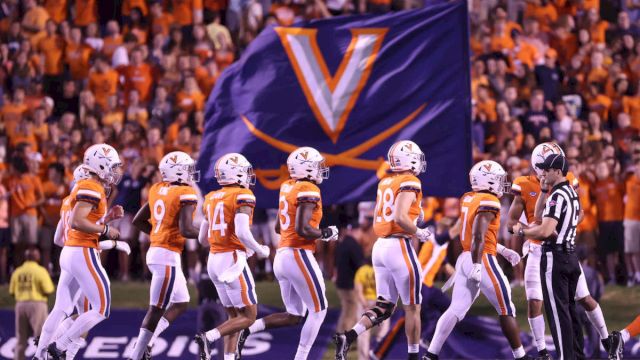 A college football team in orange and white uniforms lines up on the field as a large Virginia banner looms in the background, cheering fans abound.