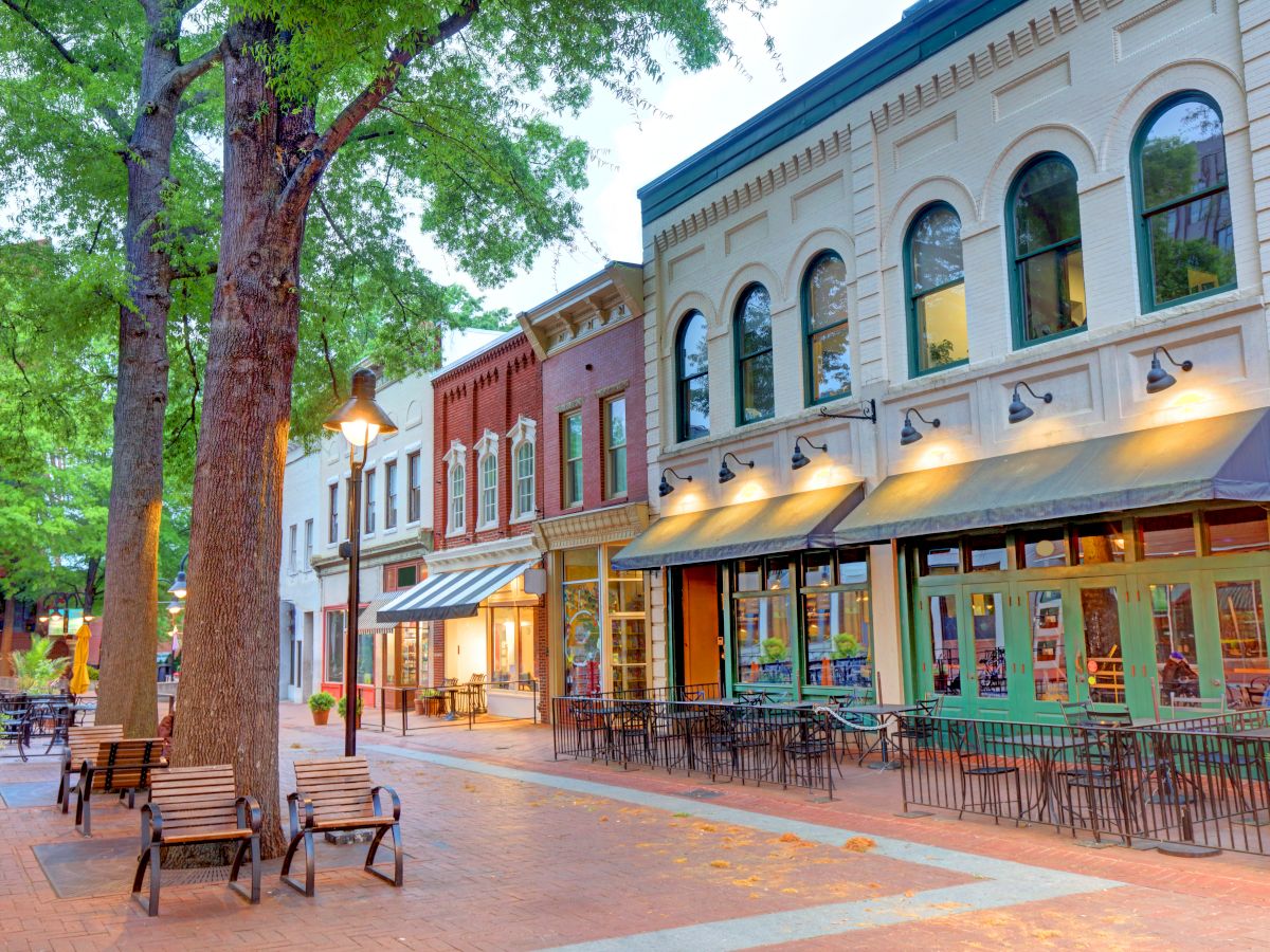 A charming town street with brick storefronts, outdoor seating, trees, and warm lights glowing at dusk.