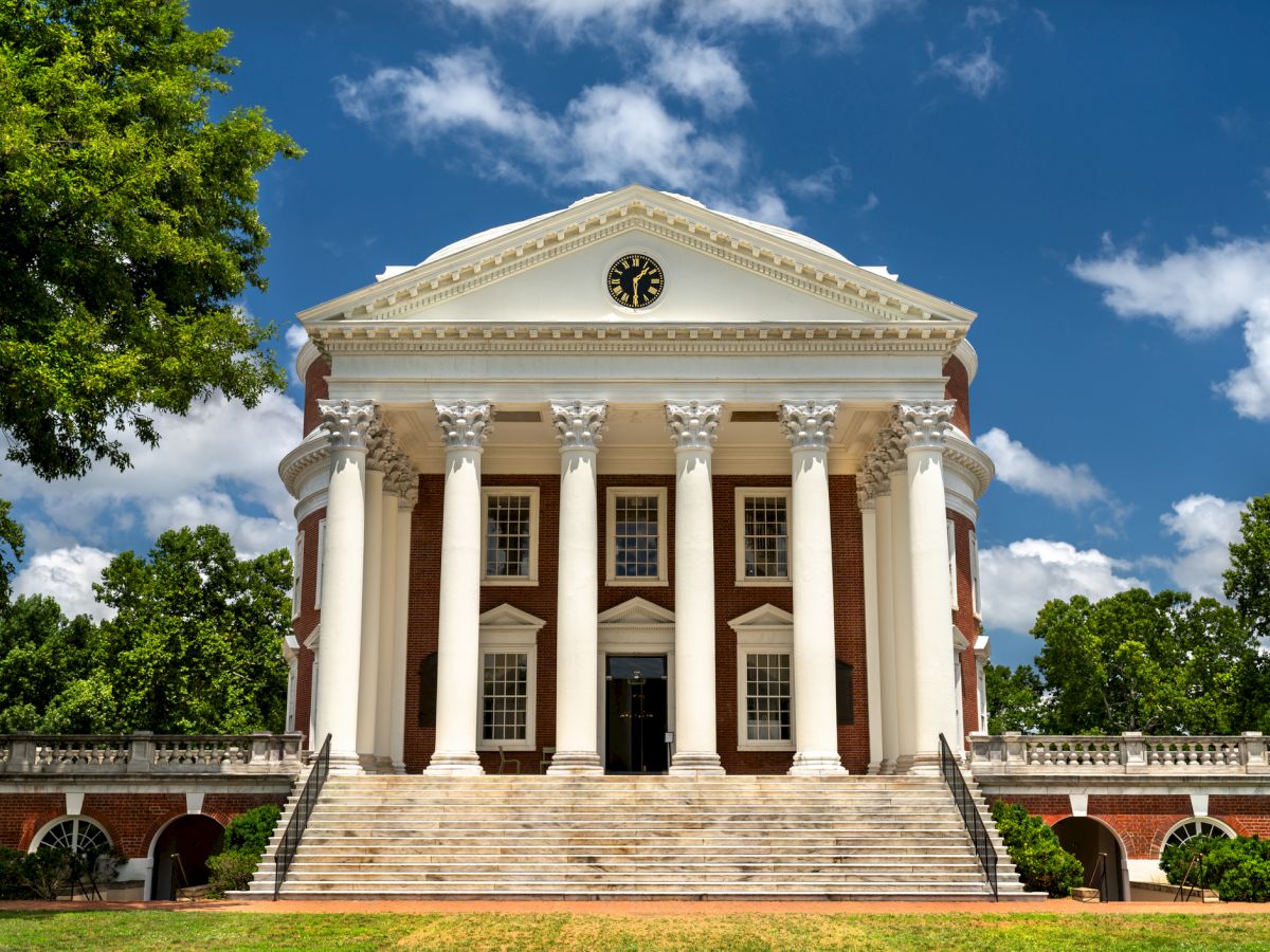 A grand white neoclassical building with tall columns, a triangular pediment, and wide steps, set in a sunny park with trees.