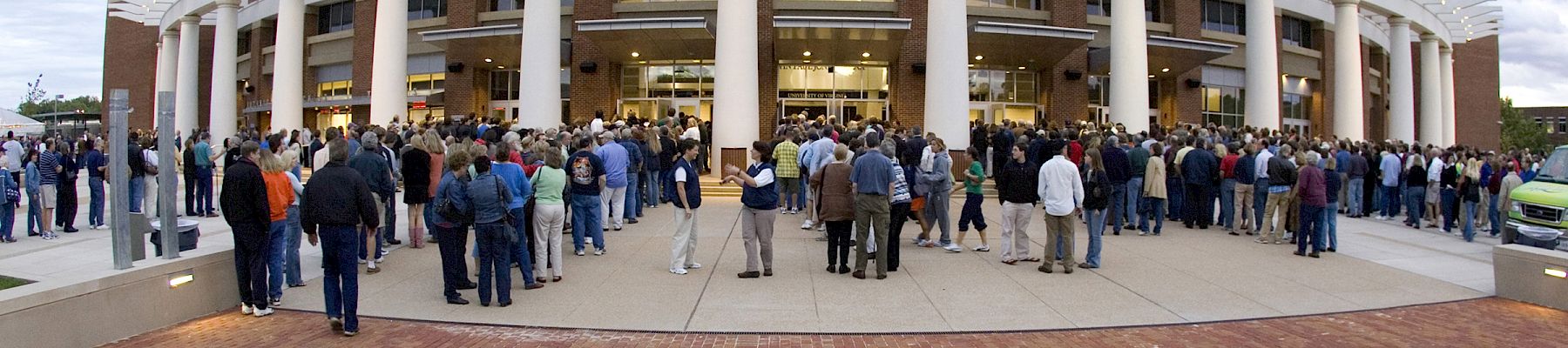 A large crowd gathers outside a modern stadium or arena with tall white pillars and a curved roof, waiting to enter.