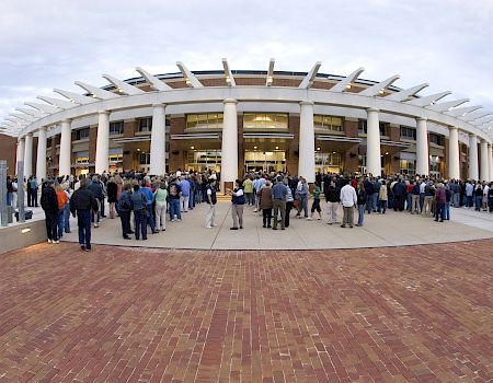 A large crowd gathers outside a modern stadium or arena with tall white pillars and a curved roof, waiting to enter.