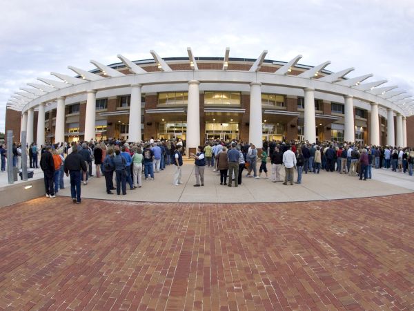 A large crowd gathers outside a circular building with tall white columns and a unique spoked roof, likely for an event or opening, everyone waiting in line.