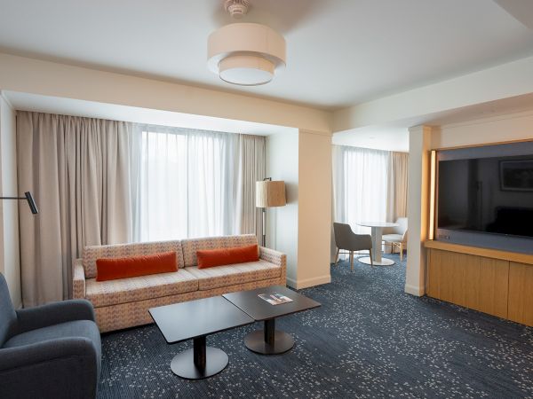 A modern hotel living area with a beige sofa, red throw pillows, blue chairs, a black coffee table, and a large TV wall, plus a bright, windowed backdrop.