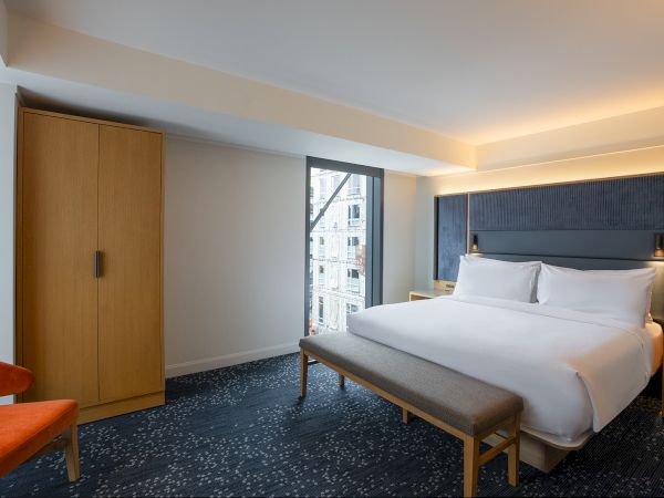 A modern hotel room with a large bed, a padded headboard, nightstands, a wooden wardrobe, a bench at the foot, a window view, and soft ambient lighting.