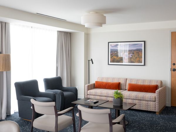 A bright hotel living area with a beige sofa and orange cushions, two dark armchairs, a glass coffee table, and a few chairs around the room.