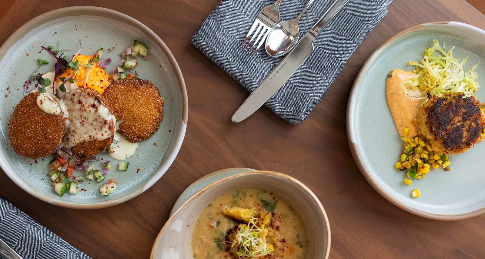 A top-down view of three plates with mixed items: breaded croquettes, salad bits, and a creamy soup or dip, plus cutlery and napkins on a wooden table.