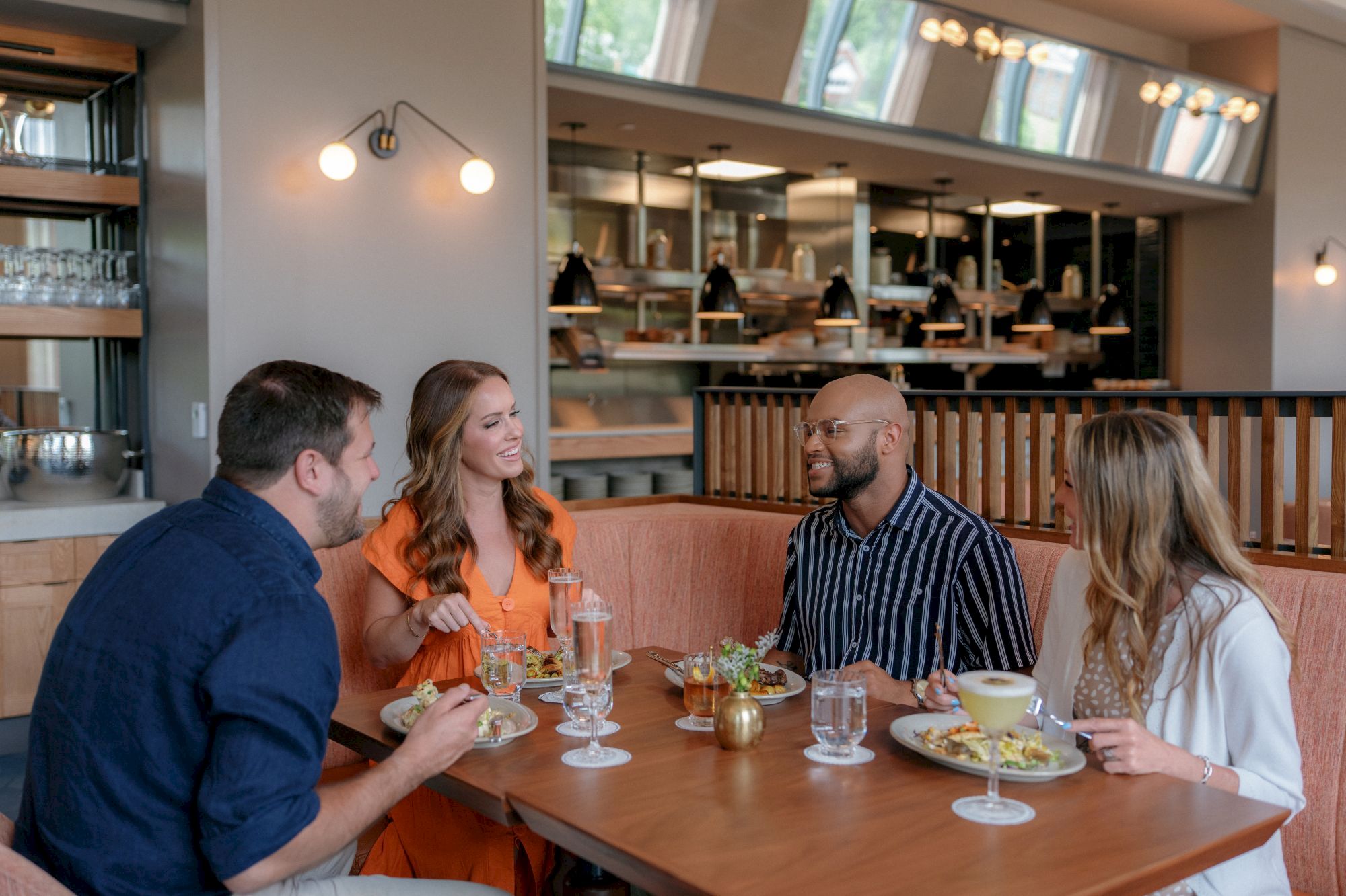 Friends enjoy a meal and drinks at a restaurant table with a lively bar in the background.