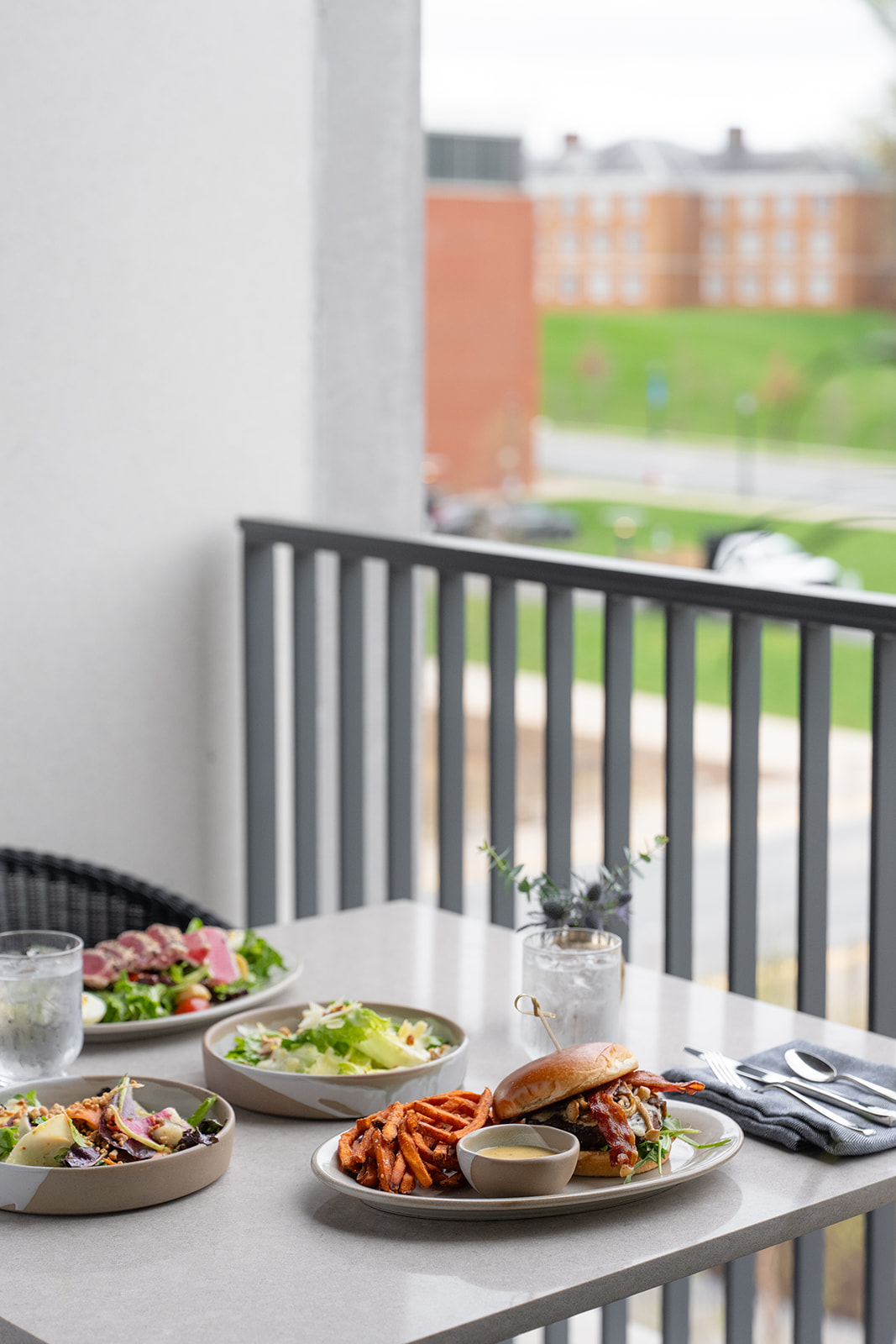 A balcony meal: four plates of salad and pasta, a small bowl of dip, drinks, and a view of buildings and a green courtyard in the background.