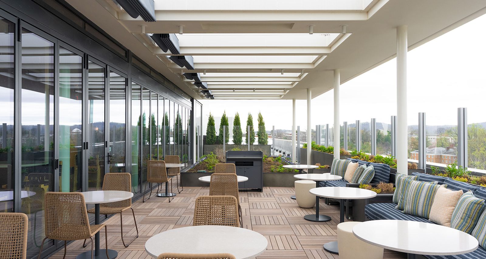 Outdoor rooftop lounge with wicker chairs, round white tables, cushioned seating, and a long row of potted evergreens under a glass canopy.