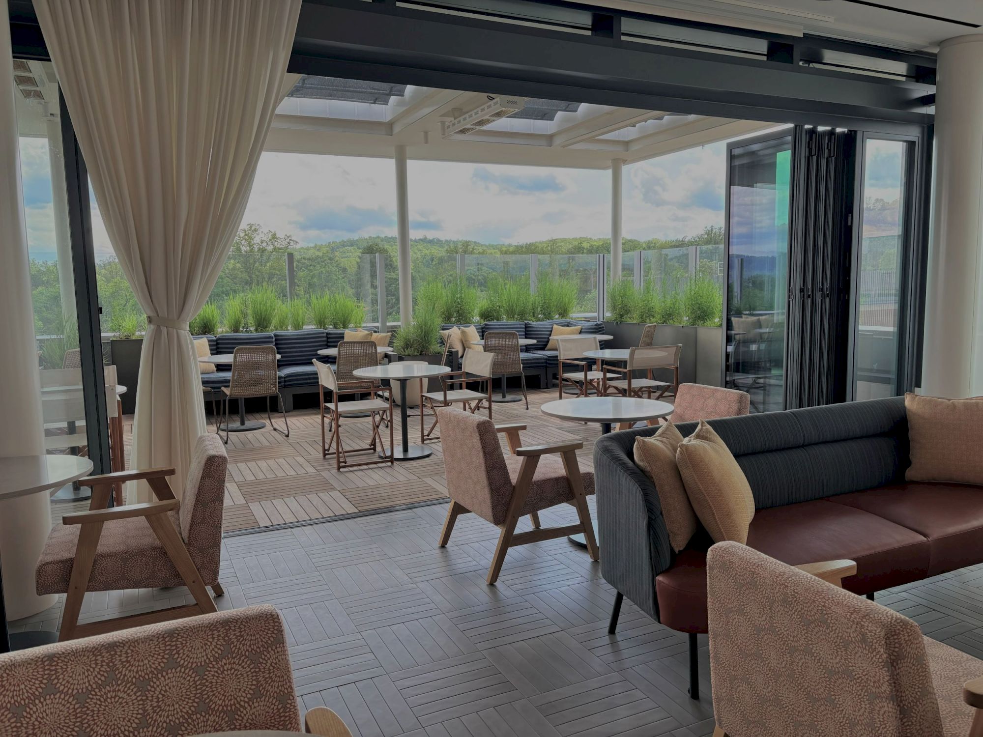 A bright hotel-style patio with cushioned sofa, chairs and round tables, overlooking green trees through glass walls.