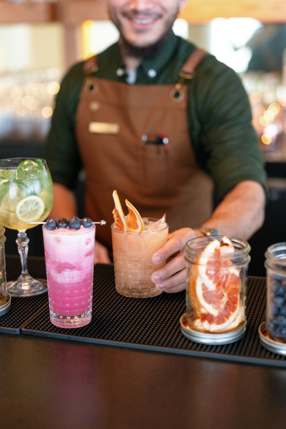 A bartender smiles behind a bar, holding an orange cocktail while colorful drinks sit on the table.
