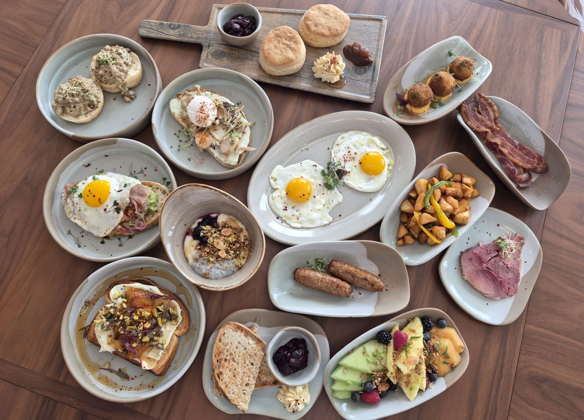 A lush breakfast/brunch spread with eggs, toast, pastries, fruit, yogurt, granola, bacon/sausages, berries, and assorted toppings on platters.