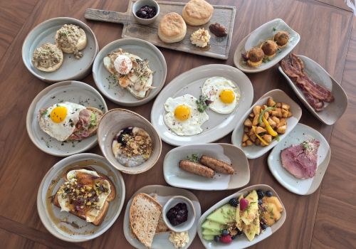 A spread of brunch plates: sunny-side-up eggs, yogurt with granola, toasted bread, pastries, fruit, sausages, bacon bits, croissants, nuts, berries, and small veggie plates, all on gray platters.