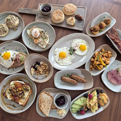 A spread of brunch plates: sunny-side-up eggs, yogurt with granola, toasted bread, pastries, fruit, sausages, bacon bits, croissants, nuts, berries, and small veggie plates, all on gray platters.